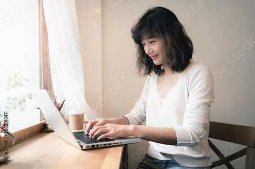 Young Asian woman working and video conferencing with laptop computer. A happy woman with smiley face working from home. A cup of coffee on wooden table. Social distancing concept