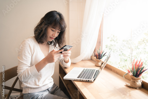 Young Asian woman using smartphone for chatting while working with laptop computer. A woman comunicating with another people while working from home. Social distancing concept photo