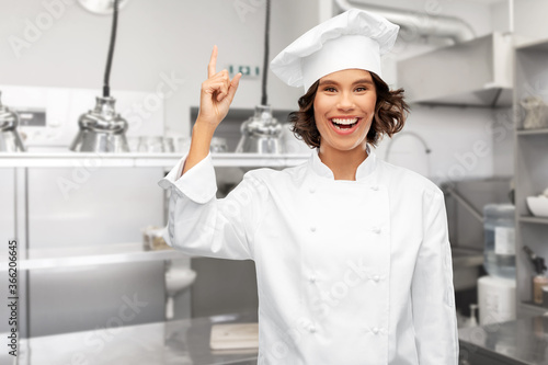 cooking, advertisement and culinary concept - happy smiling female chef in toque pointing finger up over restaurant kitchen background