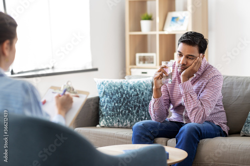 psychology, mental therapy and people concept - sad young indian man patient with glass of water and woman psychologist at psychotherapy session