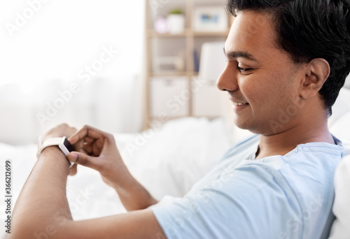 technology, bedtime and rest concept - happy smiling indian man with smart watch lying in bed at home