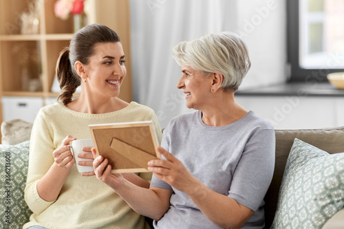 family, generation and people concept - happy smiling adult daughter and senior mother with at photo frame talking at home