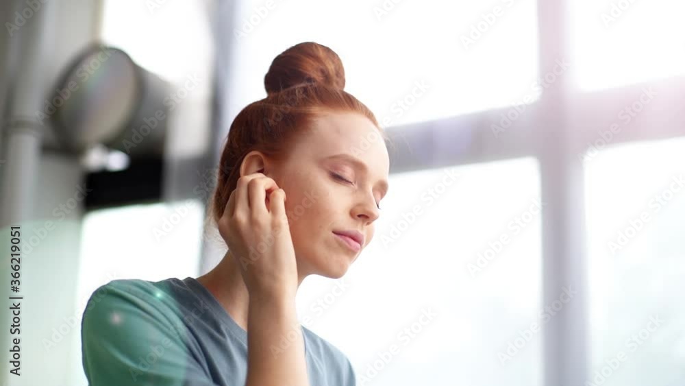 Close-up of face with emotion of joy of cheerful redhead young woman listening music in wireless earphones. Happy female having fun while listening music with closed eyes on background of large window Close-up of face with emotion of joy of cheerful redhead young woman listening music in wireless earphones. Happy female having fun while listening music with closed eyes on background of large window
