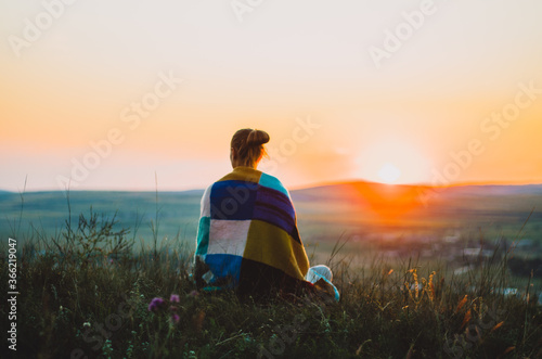 Back view of a young woman wrapped in a colourful knitted blanket seated on a hill at sunset, meditating, contemplating
