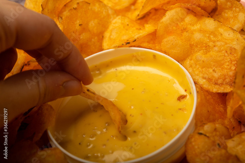 The girl takes a chip from a round dish with potato chips and a saucepan with cheese sauce in the center of the plate. Close up.