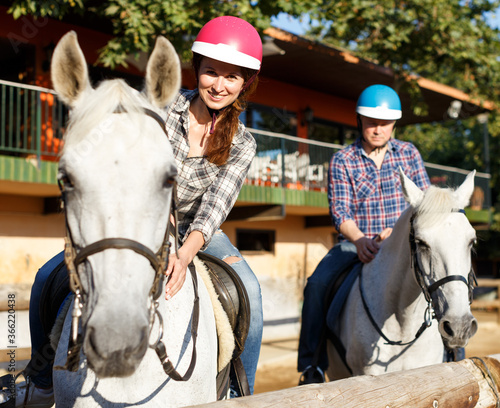 Portrait of mature couple in helmets riding by horse at barn at summer day