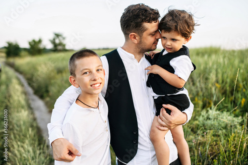 Proud father with two sons walk outdoors. Dad with toddler in his arms and preteen son stand in the scenic field