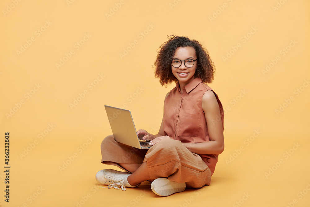 Portrait of smiling Afro-American student girl with curly hair sitting with crossed legs and using laptop against yellow background Portrait of smiling Afro-American student girl with curly hair sitting with crossed legs and using laptop against yellow background