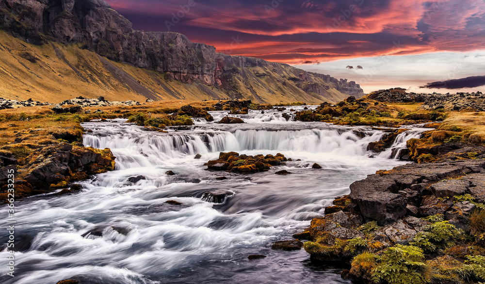 Fototapeta premium Wonderful nature of Iceland. fresh green grass and icelandic moss near river with waterfall. Tipical Icelandic scenery during sunset. Dramatic Scene of powerfull Waterfall with colorful sky