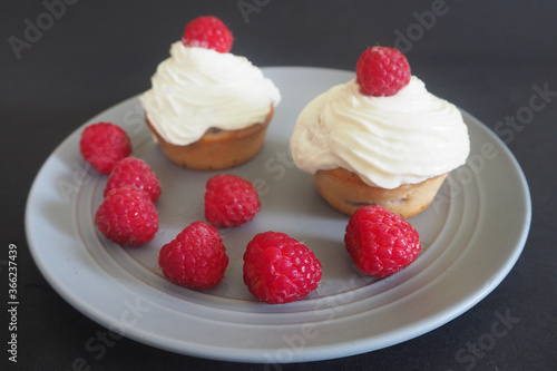 lots of raspberries on the background of two gluten-free cupcakes with cherries and mascarpone cream on a round gray plate . gluten-free homemade cakes