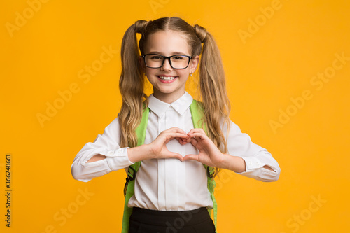 Canvastavla Smiling schoolgirl gesturing heart shape with fingers on yellow background