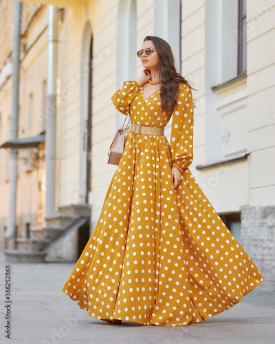 Pretty stylish woman wearing yellow and white polka-dot sundress and sunglasses. Beautiful girl in long dress walking and standing at city street © Dmitry Tsvetkov