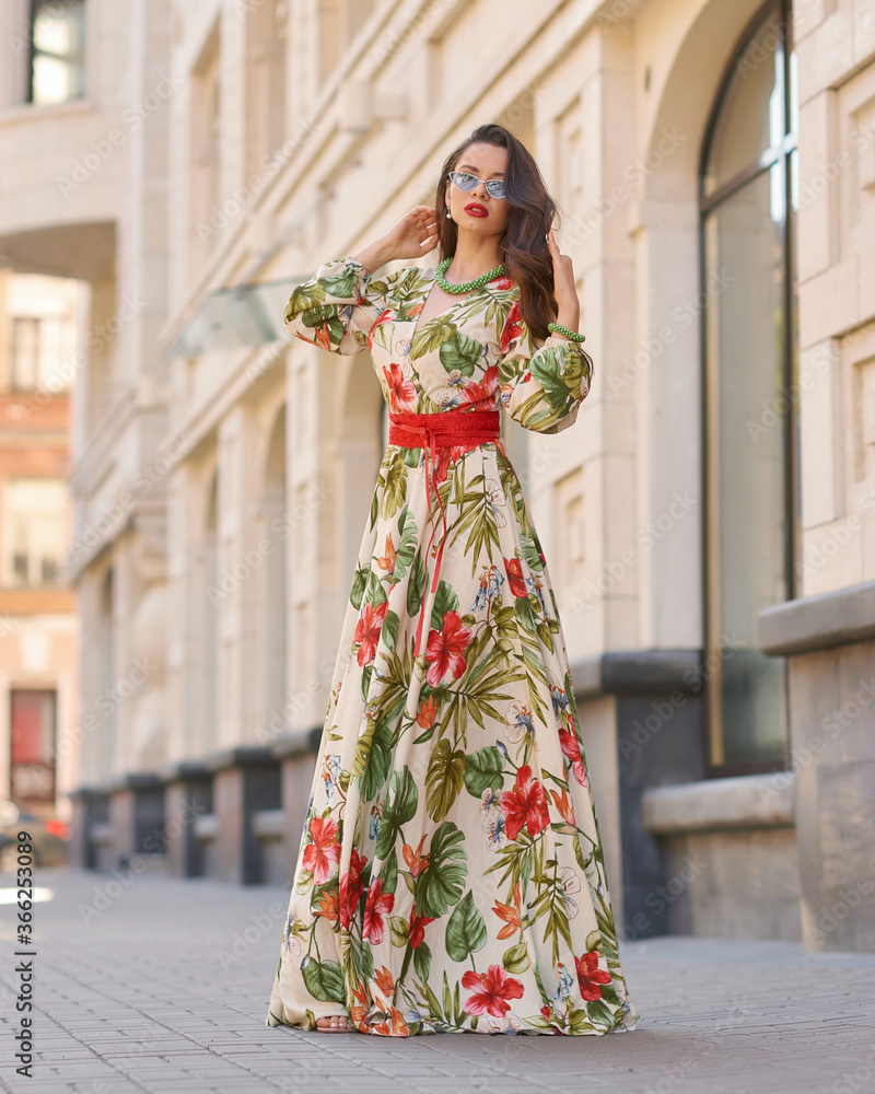 Fashion outdoor portrait. Elegant woman in colorful white red and green dress with floral design walking city street. Female model with long brunette wavy hair