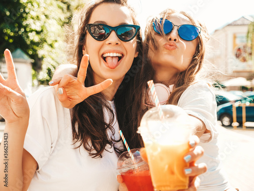 Two young beautiful smiling hipster women in trendy summer clothes. Carefree women posing outdoors.Positive models holding and drinking fresh cocktail smoothie drink in plastic cup with straw