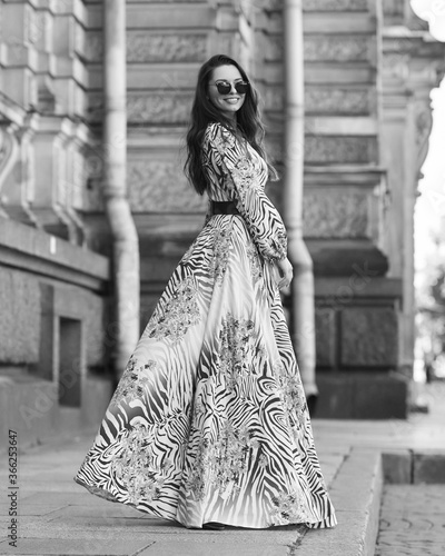 Young beautiful pretty caucasian young woman with brunette wavy hair wearing long gray summer dress and walking at city street on a summer day