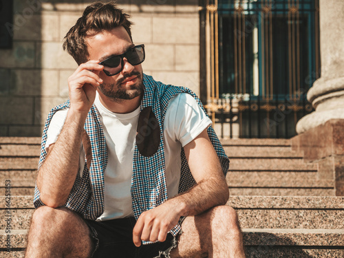 Portrait of handsome confident stylish hipster lambersexual model.Man dressed in white T-shirt. Fashion male sitting at the stairs on the street background in sunglasses photo