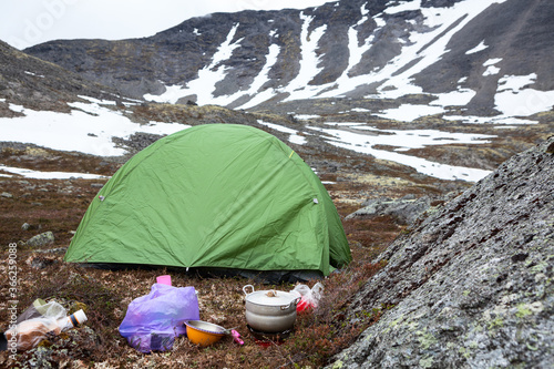 Food cooking on portable gas stove during hiking, small green tent is in mountain valley for resting, mountains with severe weather and snow patches