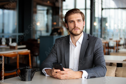 Focused mature businessman in a stylish suit and white shirt sits in a cafe with a phone in his hand and look aside 