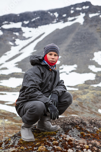 Caucasian teenage boy dressing windproof and waterproof hiking outfit for mountaineering, young man sitting on rock and looking at camera in mountains