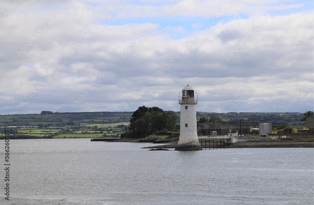 The small, white harbour lighthouse on the Shannon Estuary with the landscape of County Kerry, Ireland in the background.