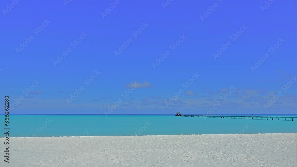 Maldivian minimalism. White sand beach, aquamarine ocean, azure sky. Above the water there is a wooden footpath and a canopy in the distance.
