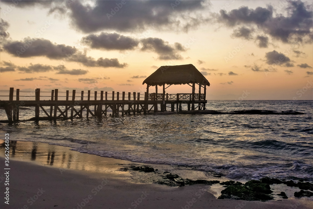 Mysterious sunrise over the Caribbean Sea. Waves roll onto the smooth sandy beach. Above the water there is a wooden footpath, a canopy. The sky is orange and pink, lilac clouds. Mexican fairy tale.