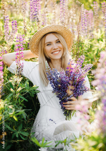 Portrait of a pretty blonde woman in a straw hat, a girl with a bouquet of lupines in her hands posing in the field.