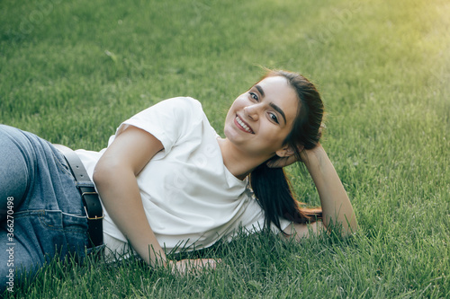 One young European brunette with dark eyes in a white T-shirt is lying on the grass holding her head and smiling. Caucasian woman laughing while lying on the lawn on a summer day. Place for text.