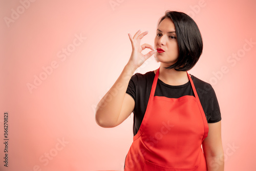 Woman employed at supermarket with red apron apron and black t-shirt showing ok