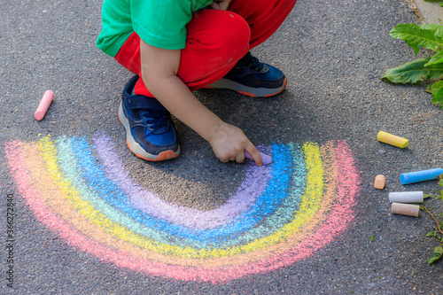 A boy draws a rainbow on the asphalt with crayons . A child draws on the asphalt . Children's drawing. Walking in the city with a child. Summer in the city. Crayons for drawing.