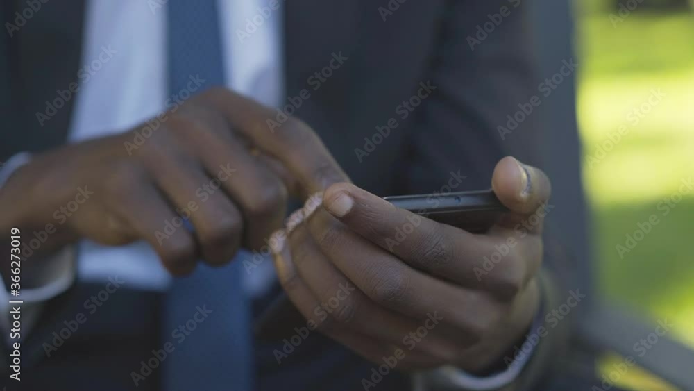 Close-up of African American male hands using smartphone. Unrecognizable businessman messaging online in wireless chat. Unknown young man sitting on bench in sunny summer park. Close-up of African American male hands using smartphone. Unrecognizable businessman messaging online in wireless chat. Unknown young man sitting on bench in sunny summer park.