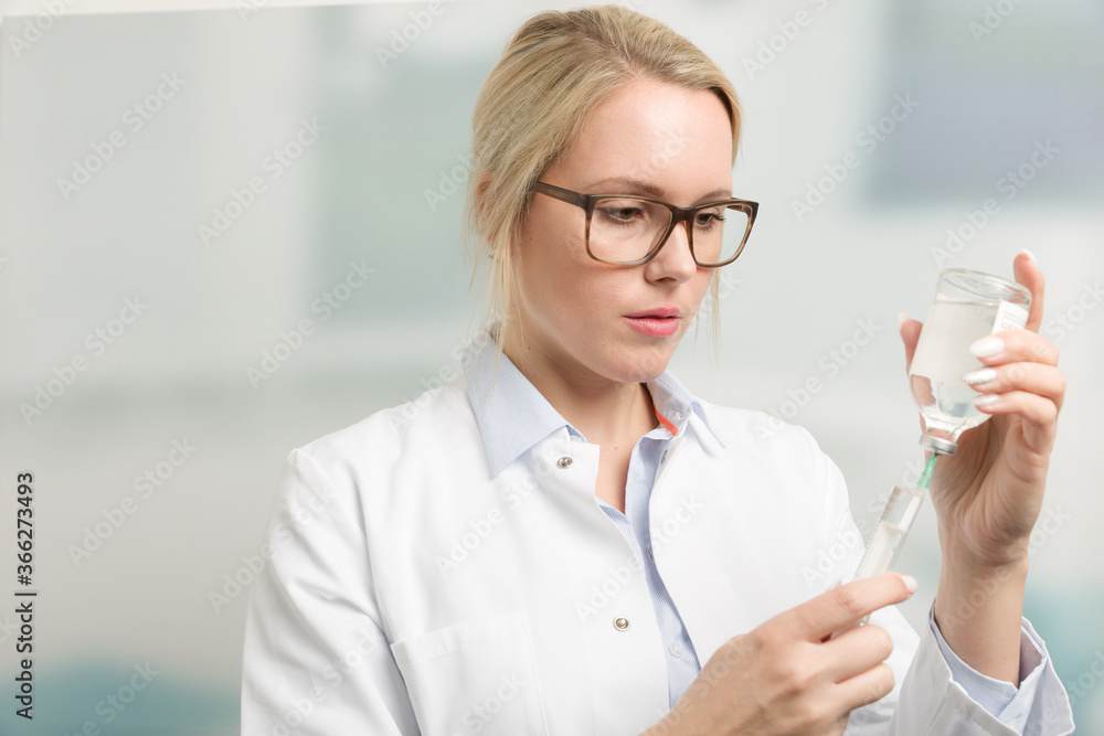 young blonde female doctor in doctor's overall is pulling up a syringe in front of a clinic room young blonde female doctor in doctor's overall is pulling up a syringe in front of a clinic room