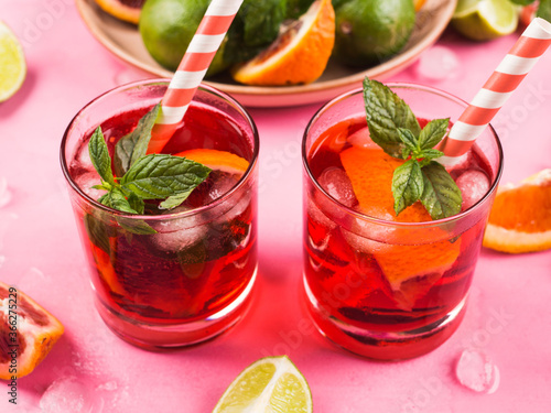 Fresh iced red cocktail in pink glasses with blood orange and lime on pink background garnished with mint leaves. Closeup
