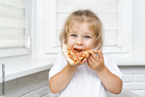 a child happily bites a piece of pizza while sitting at the table in a white kitchen