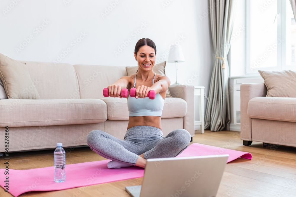 Young woman exercise with dumbbells at home and watching tutorial lesson on laptop computer. happy young female doing arm workout using dumbbells and looking at tutorial on her laptop Young woman exercise with dumbbells at home and watching tutorial lesson on laptop computer. happy young female doing arm workout using dumbbells and looking at tutorial on her laptop