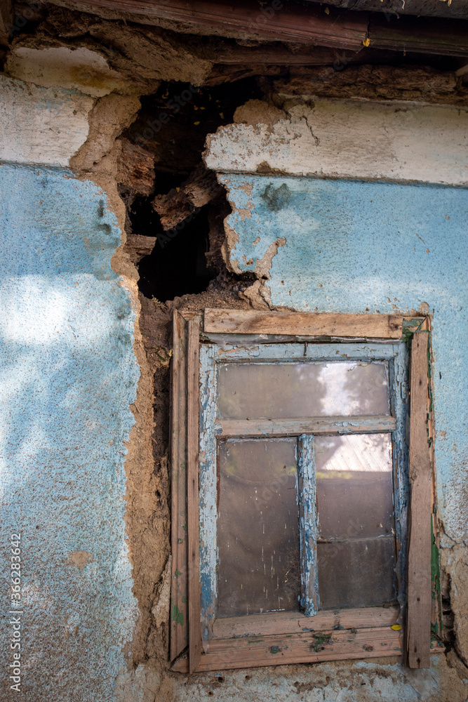 Blue painted house wall with a blue wooden window and a big crack in the wall. Hole. Blue painted house wall with a blue wooden window and a big crack in the wall. Hole.