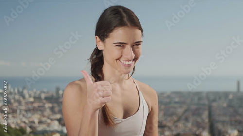 Sportswoman showing thumbs up sign. Fitness woman looking at camera outside