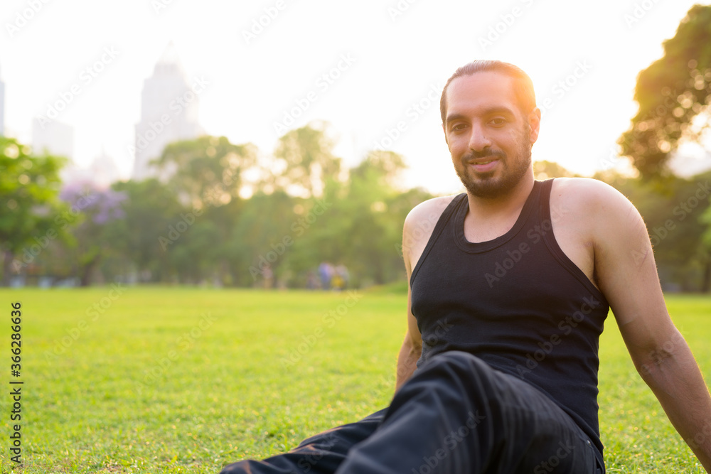 Happy young handsome bearded Indian man sitting at the park