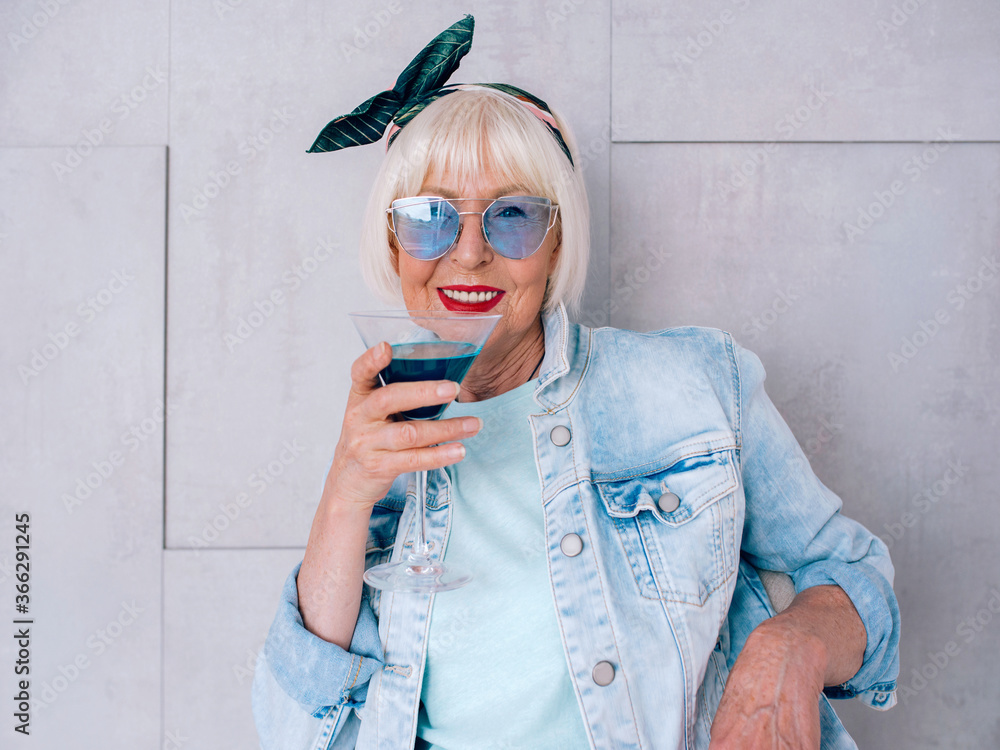 Fototapeta premium senior (old) stylish woman with gray hair and in blue glasses and jeans jacket holding glass with blue cocktail. Alcohol, relax, holidays, retirement concept