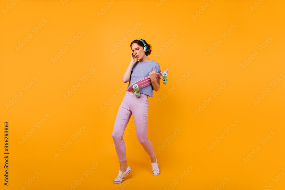 Full-length photo of dancing glamorous lady with longboard. Studio portrait of amazing caucasian woman posing in headphones and listening music.
