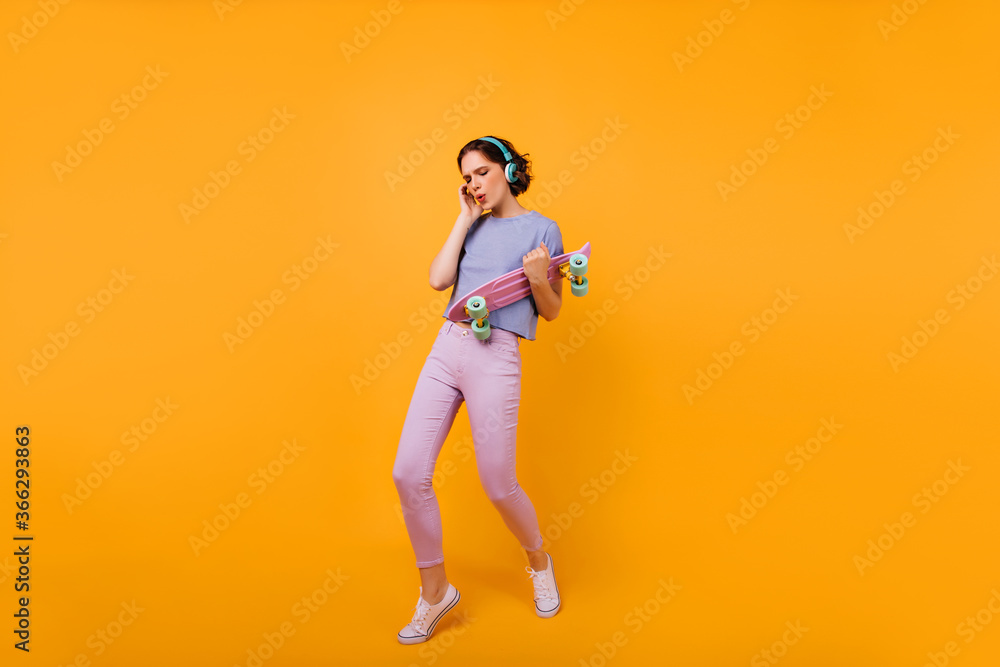 Full-length photo of dancing glamorous lady with longboard. Studio portrait of amazing caucasian woman posing in headphones and listening music. Full-length photo of dancing glamorous lady with longboard. Studio portrait of amazing caucasian woman posing in headphones and listening music.