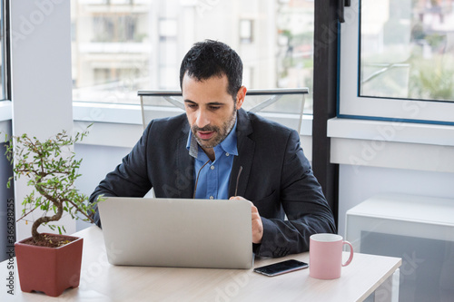 manager  vestito con giacca nera e camicia blu, seduto nella sua postazione di lavoro si concentra nello scrivere qualcosa nella tastiera del suo portatile photo