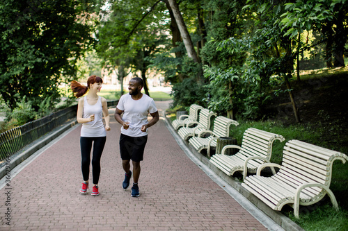 Happy joyful young multiethnic couple  African American man and Caucasian woman  jogging across the city park alley at early morning with sunrise and trees in background
