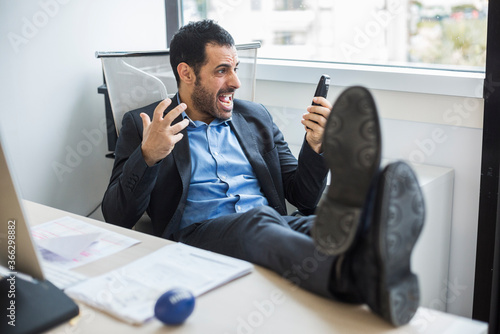 Fotografía manager moro vestito con giacca nera e camicia blu, seduto nella sua postazione di lavoro tiene i piedi sulla scrivania e si stressa dopo una telefonata