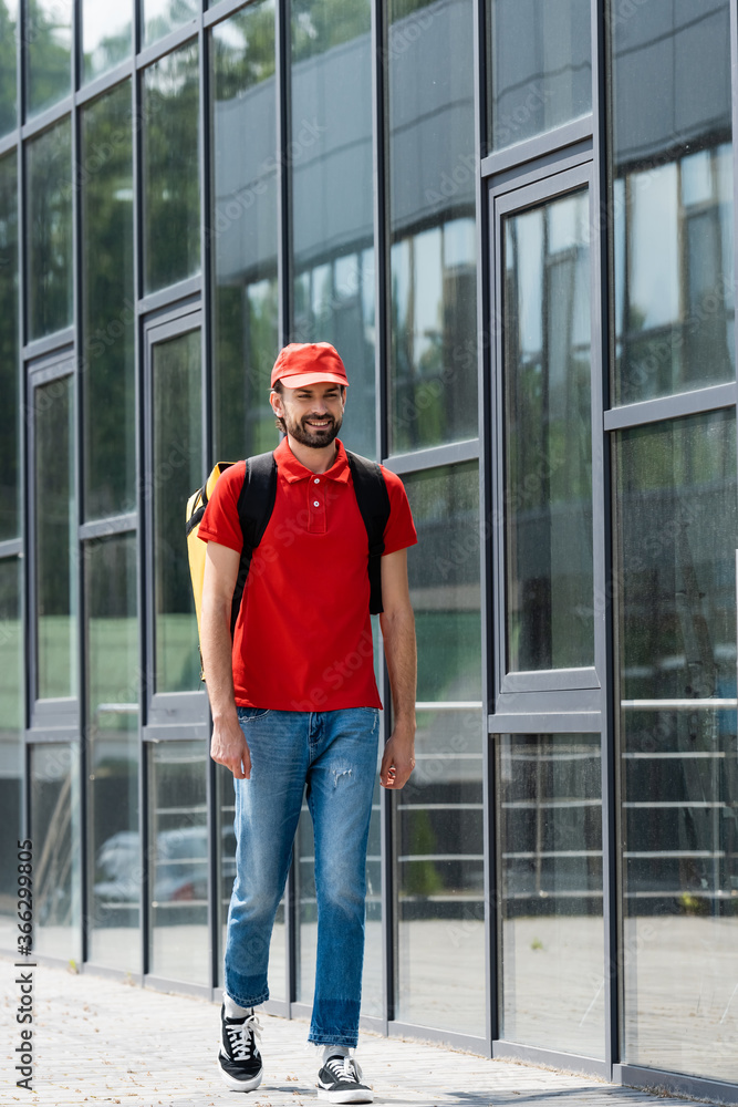 Smiling delivery man in uniform and thermo backpack walking on urban street