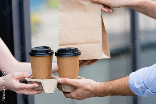 Cropped view of waitress giving coffee to go and paper bag to customer near cafe on urban street