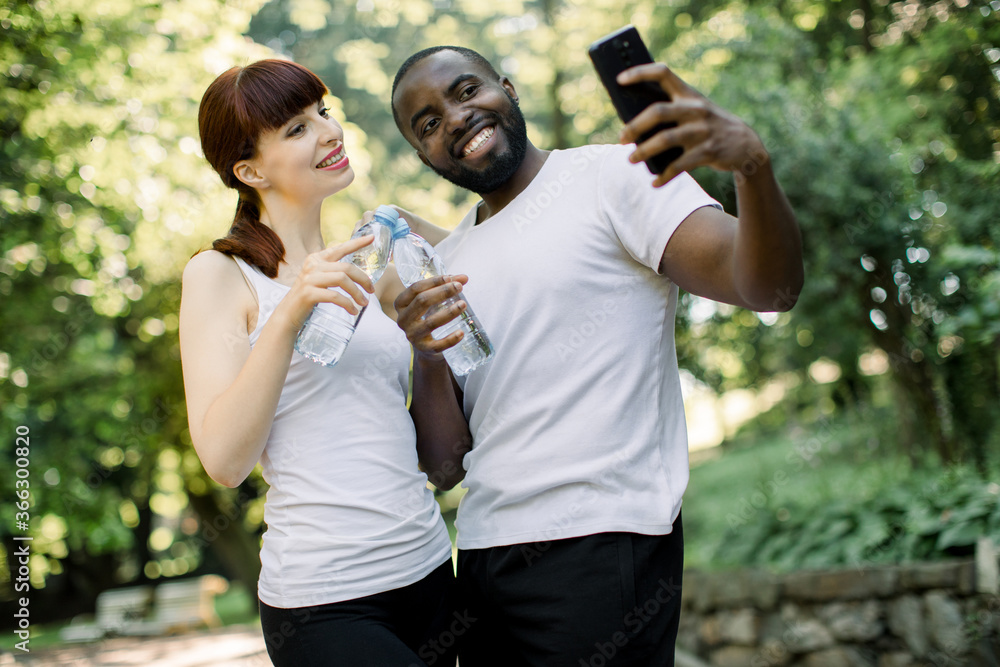 Modern joyful multiethnic friends couple with water bottles, making pause for selfie photo on smartphone, standing in urban green park during sport training, jogging or fitness