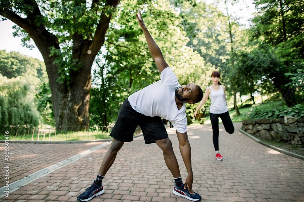 Obraz premium Sport, fitness and jogging concept. Young multietnic couple ready to run on fresh morning in the city park, stratching before exercises. Focus on African man with arm raised. Woman on the background
