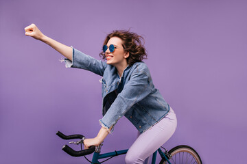 Confident girl in denim jacket riding on bike and waving hand. Indoor photo of inspired young lady in glasses sitting on bicycle.
