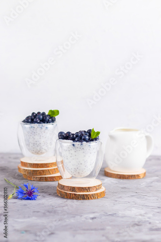 Chia pudding with mint and blueberries in glasses on a wooden stand with blue chicory flower and white cream jug on a white background